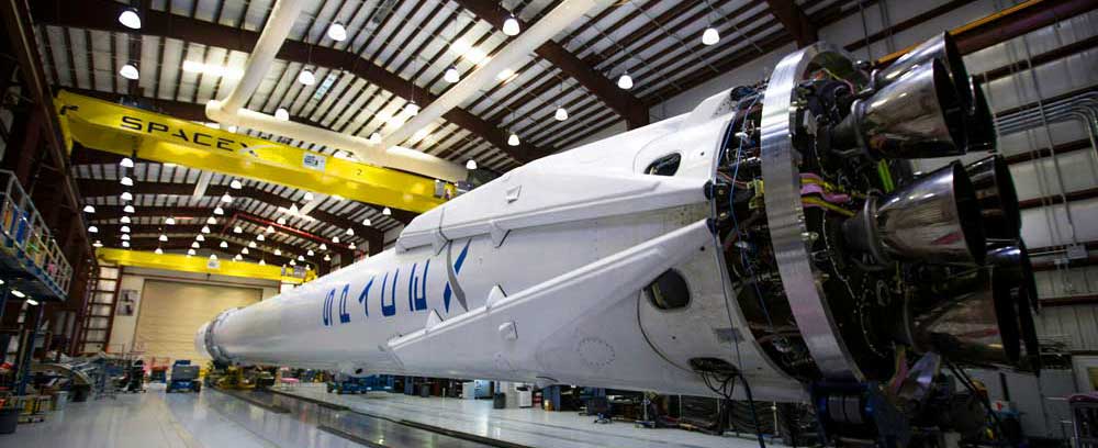 McCollisters A SpaceX rocket lies horizontally inside a large industrial facility, with its engines visible in the foreground and overhead cranes and beams above. The word SpaceX is printed on the rockets side.