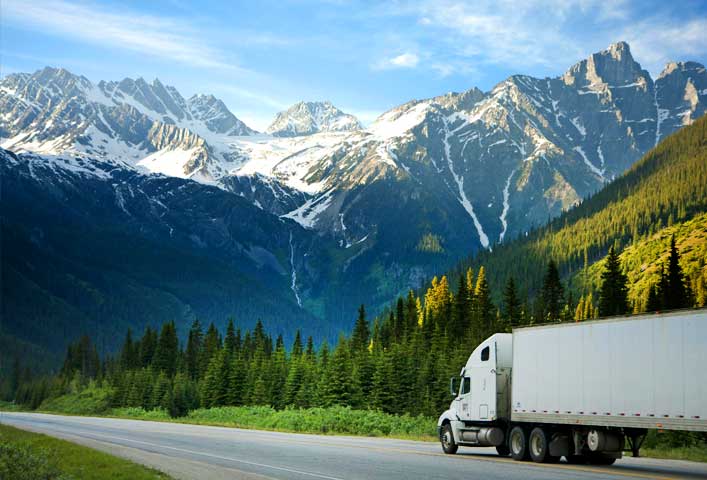 McCollisters A white semi-truck drives along a highway bordered by green trees, showcasing efficient transportation against a backdrop of snow-capped mountains and a blue sky.