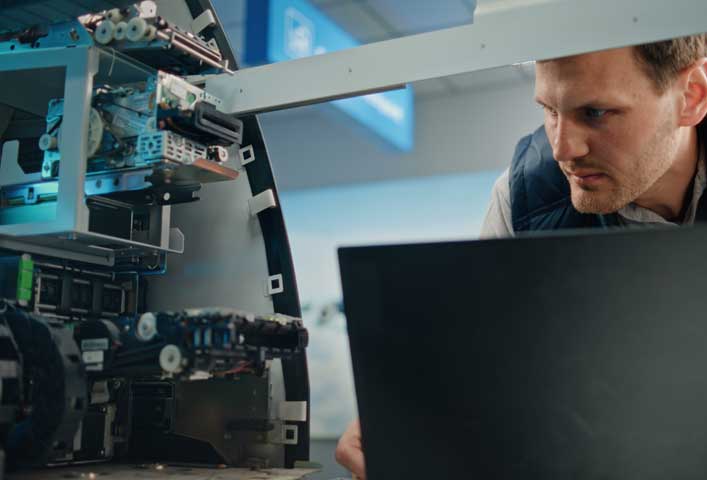 McCollisters A man closely examines machinery or equipment with exposed wiring and components, possibly during a banking installation, while working on a laptop. The background is out of focus, suggesting a technical or industrial setting.