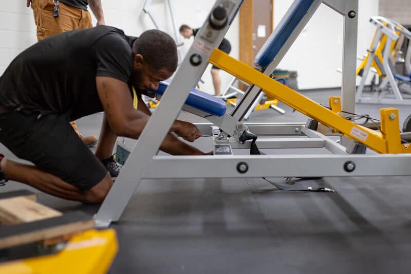 McCollisters A person kneels on the floor while handling the installation or repair of fitness equipment in a fitness center. Gym equipment and another person standing are visible in the background.
