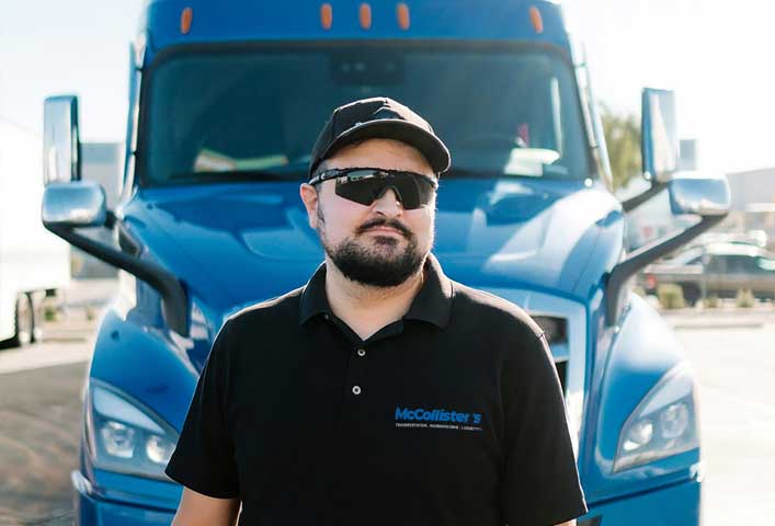 McCollisters A man wearing a black cap, sunglasses, and a black McCollisters polo shirt stands in front of a large blue Auto Transport semi-truck, outdoors on a sunny day.