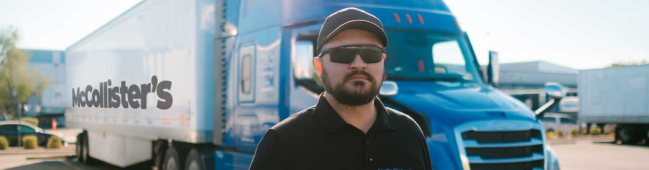 McCollisters A man wearing sunglasses and a cap stands in front of a blue semi-truck with a trailer that has “McCallister’s” written on the side. It appears to be a sunny day at a trucking facility.