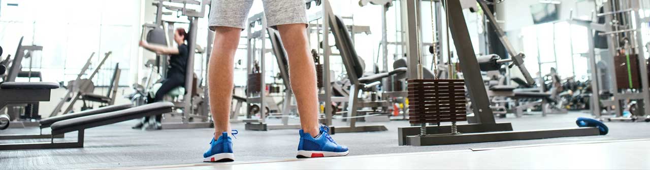 McCollisters A person in blue athletic shoes and shorts stands in a bright gym, surrounded by various exercise machines and equipment. Other people are seen working out in the background.