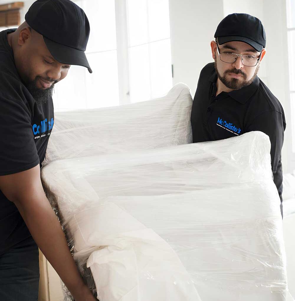 McCollisters Two movers wearing black uniforms and caps are carrying a large, plastic-wrapped piece of furniture into a bright room, ensuring your residential relocation is smooth and efficient.