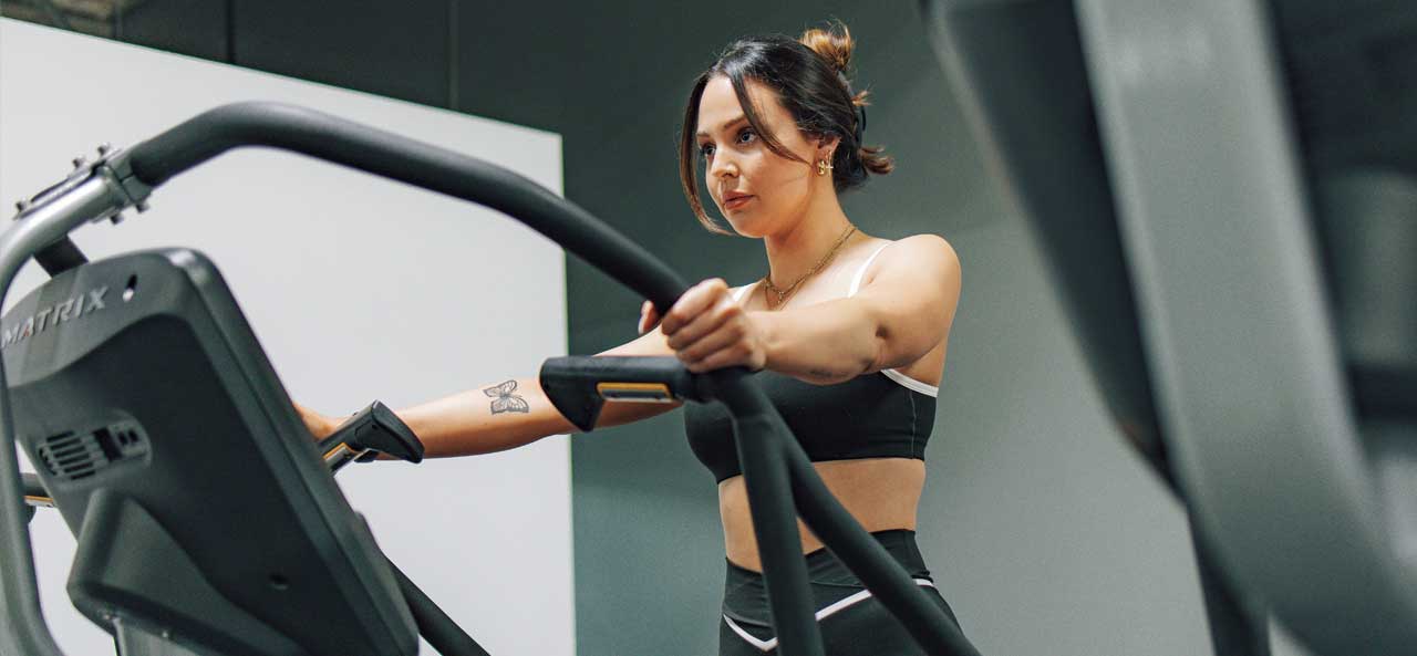 McCollisters A woman in athletic wear exercises on an elliptical machine in a gym, holding the handles and looking focused, surrounded by well-designed fitness spaces.