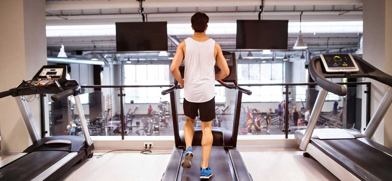 McCollisters A man wearing a white tank top and black shorts runs on a treadmill in sleek fitness spaces of a modern gym, with other equipment and people visible in the background through large windows.