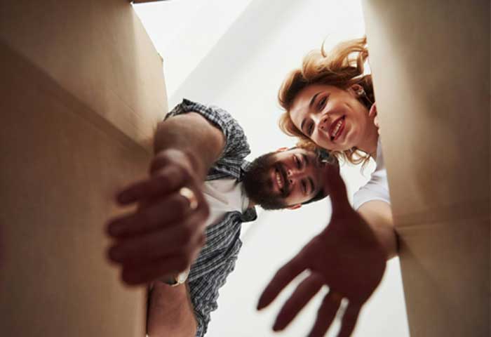 McCollisters A man and woman smiling and reaching down, as seen from inside a cardboard box, suggesting they are unpacking or packing together during a residential relocation or home moving experience.