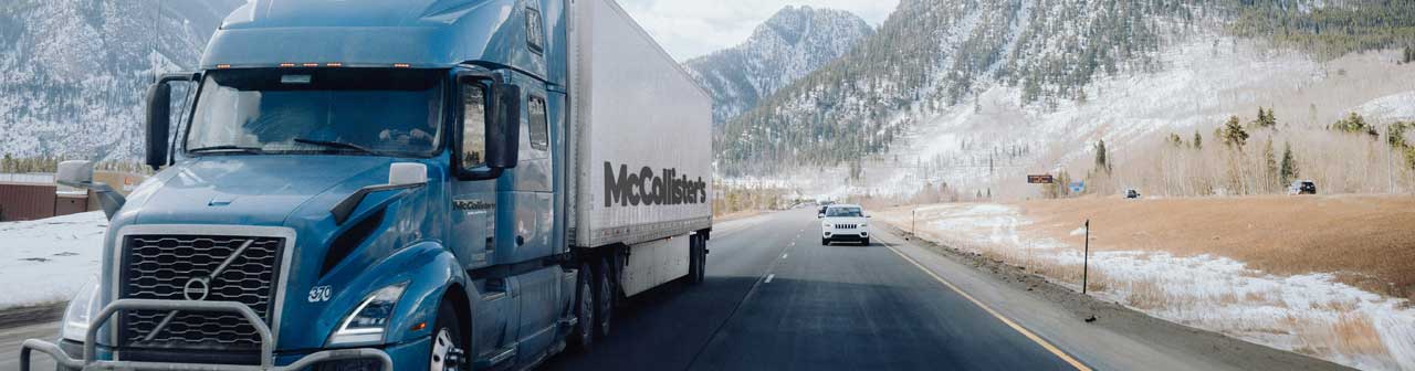 McCollisters A blue semi-truck with a white trailer, part of the vital transportation network, drives on a highway through a snowy mountainous landscape, with a few cars visible in the opposite lane under a partly cloudy sky.