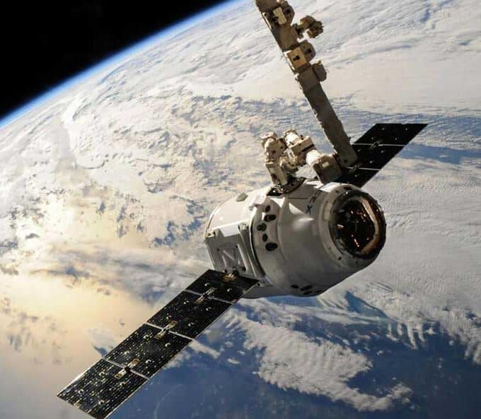 McCollisters A satellite or spacecraft, supported by an aerospace logistics partner, orbits above Earth with solar panels extended and the planet’s cloud-covered surface visible in the background.